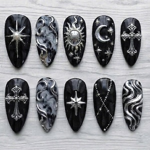 Peut inclure: Un ensemble de dix faux ongles en forme d'amande avec une base noire et diverses décorations métalliques argentées, incluant une étoile avec une perle centrale, des motifs tourbillonnants, un soleil, un croissant de lune et des croix.