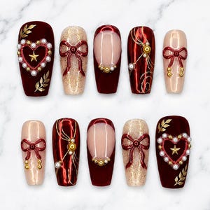 May include: A set of ten press-on nails featuring a deep burgundy and nude colour palette. Designs include 3D red bows, gold star accents, white pearl borders around heart shapes, gold leaf patterns, and metallic red finishes with gold swirls.