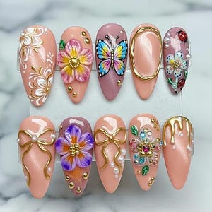 Puede incluir: Un conjunto de diez uñas artificiales con varios diseños. Las uñas presentan una gama de colores, incluyendo rosa, morado y dorado. Los diseños incluyen flores, mariposas, lazos y detalles dorados. Algunas uñas tienen elementos 3D.