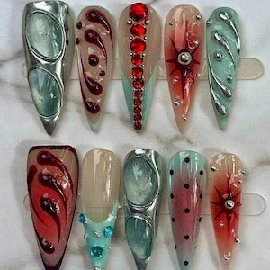 May include: A set of ten press-on nails featuring diverse designs including metallic silver accents, deep red swirl patterns, rows of bright red rhinestones, starburst motifs, light blue base colors, and small black polka dots on a glossy finish.