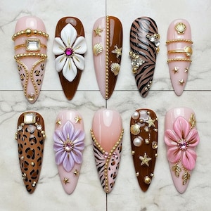 May include: A collection of ten artificial nails in various designs. The nails feature a range of colors, including pink, brown, and gold, with embellishments such as flowers, animal prints, and seashells. Each nail has a unique design.