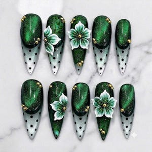 May include: A set of ten press-on nails featuring a pointed shape. The design combines a deep emerald green cat-eye effect with a translucent base adorned with black polka dots. Several nails are decorated with 3D white floral accents and gold beads.