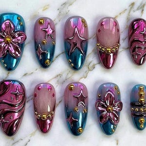May include: A collection of ten artificial nails with various designs. The nails feature a color palette of teal, pink, and gold. Some nails have flower, star, and swirl designs. Others have gold bead accents.