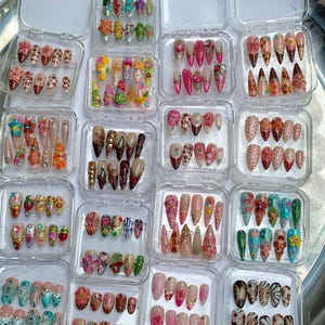 May include: A collection of sixteen clear plastic cases, each containing a set of ten press-on nails. The nails feature diverse designs including floral patterns, animal prints, rhinestones, glitter, and various colorful painted motifs.