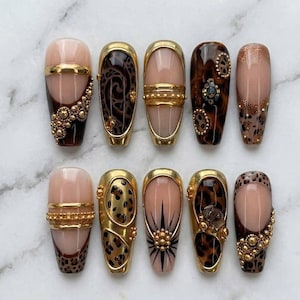 May include: A set of ten press-on nails featuring a nude base with varied designs including gold metallic accents, leopard print patterns, raised gold bead embellishments, and black starburst motifs. Each nail has a glossy finish and gold trim.