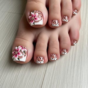 Puede incluir: Primer plano de un pie con pedicura con diseños de uñas. Los dedos gordos tienen una base blanca con diseños de flores rosas y blancas. Las otras uñas de los pies tienen una base nude con lunares negros y puntas blancas. Las uñas están sobre una superficie de madera clara.