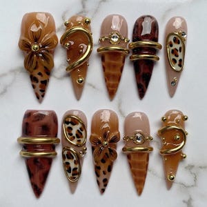 Puede incluir: Un juego de diez uñas postizas en forma de estilete con tonos marrones, bronceados y crema. Los diseños incluyen flores esculpidas en 3D, anillos de metal dorado, estampados de leopardo, pedrería transparente y pequeñas esferas doradas.