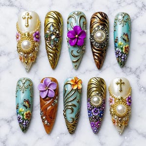May include: A collection of ten ornate, long, and pointed artificial nails. Each nail features intricate designs, including gold filigree, pearls, rhinestones, and 3D floral accents in various colors. Some nails have crosses or butterflies.