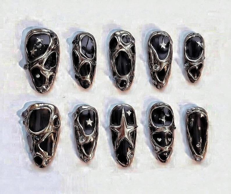 May include: A set of ten press-on nails featuring a black base with a raised, liquid-like silver metallic overlay. The design includes various cutouts, small embedded star shapes, and tiny clear rhinestones integrated into the textured silver surface.