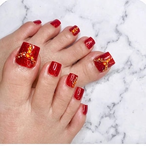 May include: Toenails painted with a glossy, glittery red polish. The large toenails on both feet feature an intricate design of small, sparkling orange and yellow rhinestones arranged in a starburst pattern, adding texture and shine to the vibrant red base.