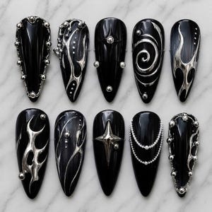 May include: A set of ten almond-shaped press-on nails in glossy black, each featuring unique 3D silver metallic accents, including raised spheres, swirling liquid-like patterns, sharp star shapes, and delicate silver beaded chains.