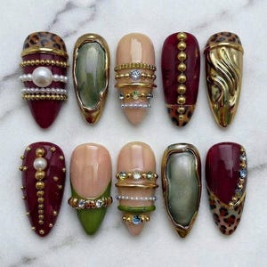 May include: A set of ten almond-shaped press-on nails featuring a variety of designs including deep burgundy, olive green, and nude base colours. The nails are decorated with gold metallic accents, small white pearls, leopard print patterns, and clear rhinestones arranged in horizontal bands and vertical lines.