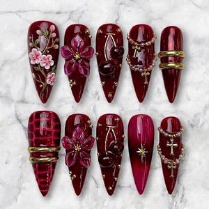 Pode incluir: Um conjunto de dez unhas postiças em formato stiletto na cor bordô profundo. Os designs incluem flores de cerejeira em 3D, flores roxas translúcidas, cerejas escuras, pingentes de cruz dourados com correntes de contas, anéis dourados e padrão de crocodilo.