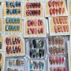 May include: A collection of press-on nail sets featuring various designs, including metallic accents, floral patterns, cross motifs, and rhinestones. The sets are displayed on individual cards labeled with sizes XS, S, M, and L, or in clear plastic cases.