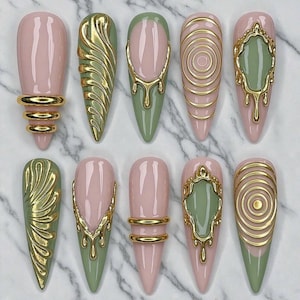 May include: A set of ten press-on nails featuring a pointed almond shape with a glossy finish. The nails are decorated in a color palette of soft pink and sage green, accented with raised, metallic gold 3D designs including rings, swirls, and drips.