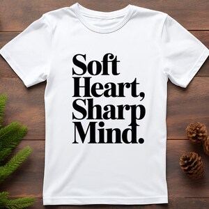 May include: White t-shirt with the text "Soft Heart, Sharp Mind" in bold black letters. The shirt is displayed on a wooden surface with pine needles and pine cones in the background.