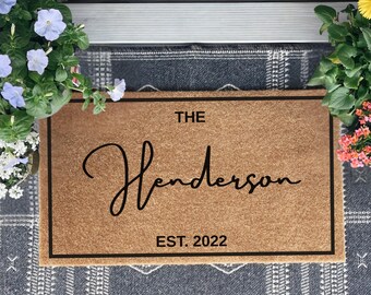May include: A rectangular doormat with the text "THE Henderson EST. 2022" in black script. The mat is brown with a black border, set on a patterned gray rug. Potted flowers with blue and yellow blooms frame the mat.