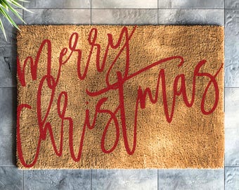 May include: A rectangular doormat made of natural fibers, with the words "Merry Christmas" in red cursive script. The mat has a light brown color and is placed on a gray tiled surface. A small potted plant is visible in the upper left corner.