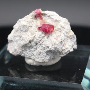 May include: A rough, light gray rock formation with two vibrant, red crystal formations. The rock sits on a clear, rectangular glass base, with a dark background. The crystals are a deep, saturated red.