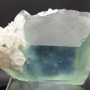 May include: A large, translucent fluorite mineral specimen featuring a prominent cubic crystal structure with a gradient of pale green to deep blue hues, accented by a cluster of smaller, opaque white crystalline formations on the left side.