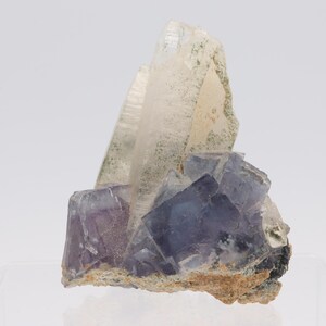 Könnte beinhalten: Ein natürliches Mineralexemplar mit einem Cluster aus lila und klaren Kristallen. Die Kristalle haben eine geometrische Form und sind auf einer beigen Matrix angeordnet. Die klaren Kristalle haben einen leichten Grünstich.