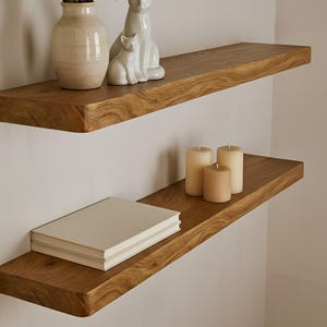 Könnte beinhalten: Zwei schwebende Holzregale an einer weißen Wand. Das obere Regal enthält eine Vase mit gelben und weißen Blumen und eine weiße Keramik-Katzenfigur. Das untere Regal hat einen Stapel Bücher und drei Kerzen.
