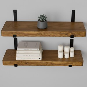 May include: Two-tier wooden floating shelves with black metal brackets. The top shelf holds a small potted succulent. The bottom shelf displays a stack of books and four white candles. The shelves are mounted on a gray wall.