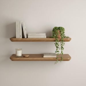 May include: Two wooden floating shelves mounted on a white wall. The top shelf holds books and a potted plant with trailing green vines. The bottom shelf has a candle, a small wooden disc, and a book. The shelves are made of light-colored wood.