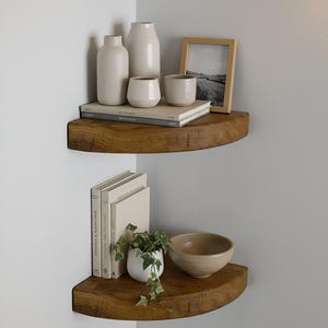 May include: Two wooden corner shelves displaying home decor. The top shelf features white vases, a small cup, books, and a framed landscape. The bottom shelf holds books, a small plant, and a beige bowl. The shelves are made of wood.