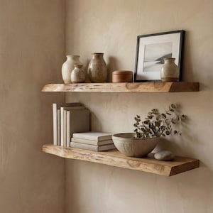 May include: Two wooden shelves mounted on a beige wall, displaying decorative items. The top shelf holds vases, a small wooden box, and a framed picture. The bottom shelf features books, a bowl with greenery, and a decorative stone.