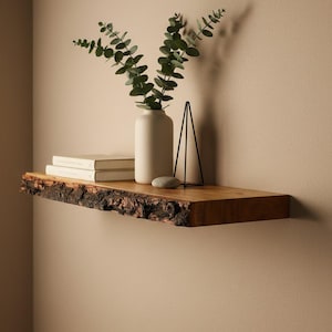 May include: A rustic wooden shelf with a live edge, mounted on a beige wall. The shelf holds a white vase with green foliage, two books, a small stone, and a black metal geometric sculpture. The shelf is approximately 30 inches long.
