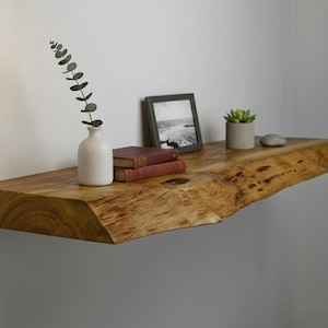 Puede incluir: Estante de madera con borde natural montado en la pared, que muestra un jarrón blanco con vegetación, dos libros, una foto enmarcada, una pequeña planta en maceta y una piedra decorativa. El estante tiene vetas de madera natural.