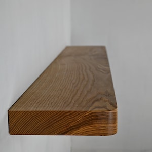 May include: A floating wooden shelf with a natural wood grain finish. The shelf is rectangular with rounded corners and is mounted on a white wall. The wood has a warm, light brown color.