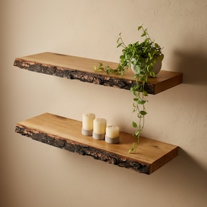 May include: Two wooden floating shelves with live edge detailing are mounted on a beige wall. The top shelf holds a small potted plant with trailing vines. The bottom shelf displays three white candles with gray bases.