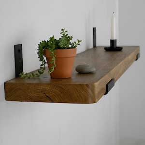 May include: A rustic wooden floating shelf with a live edge, supported by black metal brackets. The shelf holds a small terracotta pot with a trailing green plant, a gray stone, and a black candle holder with a white candle.