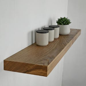 May include: A wooden floating shelf with a natural wood grain finish. Three cream-colored canisters with black lids and a small potted succulent sit on the shelf. The shelf is mounted on a white wall.