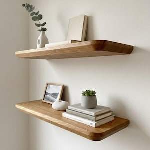 May include: Two floating wooden shelves mounted on a white wall. The top shelf holds a white vase with greenery and a book. The bottom shelf displays a framed picture, a white decorative object, books, and a small potted plant.