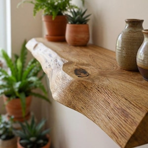 Puede incluir: Estantería de madera con borde natural, mostrando la forma orgánica de la madera. La estantería es de color marrón claro, con vetas visibles y un nudo. Pequeñas plantas en macetas y jarrones de cerámica están colocados, creando un ambiente natural y rústico.
