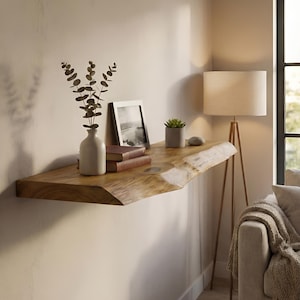 Puede incluir: Estante de madera montado en la pared con borde natural. Muestra un jarrón blanco con vegetación, una foto enmarcada, una pequeña planta en maceta y libros. Una lámpara de pie y un sofá son visibles en el fondo. El estante es de madera.