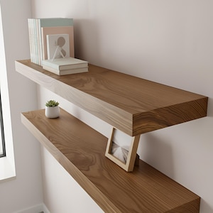 May include: Two floating wooden shelves with a natural wood grain finish are mounted on a light pink wall. The top shelf holds books and a small framed picture. The bottom shelf displays a small potted plant and a framed photo.