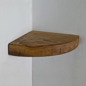 Może przedstawiać: Narożna półka z jasnobrązowego drewna. Półka ma kształt ćwierć okręgu, z zaokrągloną przednią krawędzią i płaską tylną krawędzią do montażu na ścianie. Widoczne usłojenie drewna.