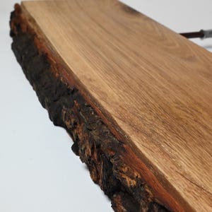 May include: A natural wood shelf with a live edge. The shelf is made of light brown wood with a dark brown, textured edge. The wood grain is visible, and the shelf appears to be unfinished. The shelf is angled, and the background is white.