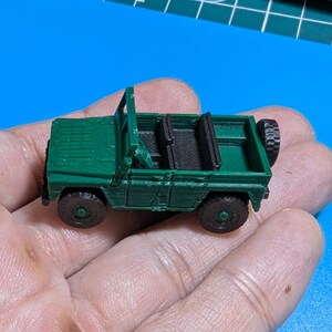 May include: A miniature green toy jeep with black wheels and interior. The jeep has a spare tire mounted on the back and a roll bar. The toy is held in a person's hand against a blue background.
