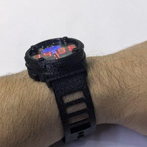 Könnte beinhalten: Schwarze Digitaluhr mit rechteckigem Zifferblatt und passendem Armband. Das Zifferblatt zeigt rot und blau beleuchtete Ziffern. Das Armband hat eine Reihe rechteckiger Aussparungen. Die Uhr wird am Handgelenk getragen.