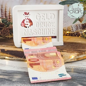 Könnte beinhalten: Eine weiße Spardose mit den Worten "Geld Druck Maschine" und einem Geldsack-Design. Zwei Euro-Banknoten kommen aus dem Schlitz. Die Banknoten sind orange, rosa und gelb.