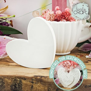 Op de afbeelding: Een wit hartvormig decoratief item staat op een houten oppervlak, naast een witte, kopvormige vaas gevuld met roze bloemen. De afbeelding bevat ook een ronde sticker met tekst en een hartontwerp.