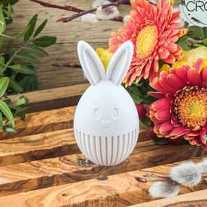 Könnte beinhalten: Ein weißes, osterhasenförmiges Dekorationsobjekt mit einem lächelnden Gesicht und vertikalen Linien am Boden. Der Hase ist vor einem Hintergrund aus bunten Blumen und Holzbrettern platziert, was ein festliches Frühlingsmotiv erzeugt.