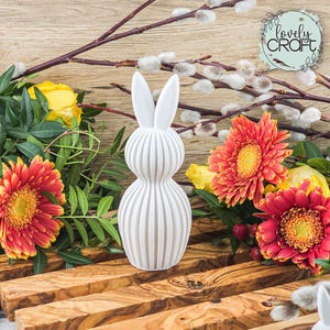Könnte beinhalten: Eine weiße, gerippte Keramik-Hasenfigur mit langen Ohren, umgeben von bunten Blumen und Grün. Das Arrangement umfasst orange und gelbe Gerbera-Gänseblümchen und Kätzchenzweige, die ein Frühlings- oder Osterthema schaffen.