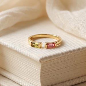 Könnte beinhalten: Ein goldener offener Ring mit zwei im Baguetteschliff geschliffenen Edelsteinen, einer in hellgrüner Farbe und der andere in einem Rosaton, platziert auf der Oberkante eines Stapels aus zwei Büchern. Der Ring hat eine polierte Metalloberfläche.
