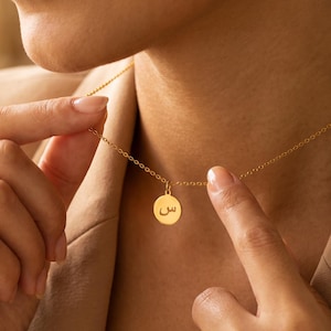 May include: A gold-toned necklace featuring a delicate chain and a small, circular pendant. The pendant is engraved with the Arabic letter seen, which is centered on the disc. The necklace is worn around the neck, showcasing the metallic finish.